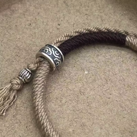 Bracelet Cercle Du Bonheur : cordon beige et noir avec fermoir argenté à motif, nœud et pendentif torsadé.